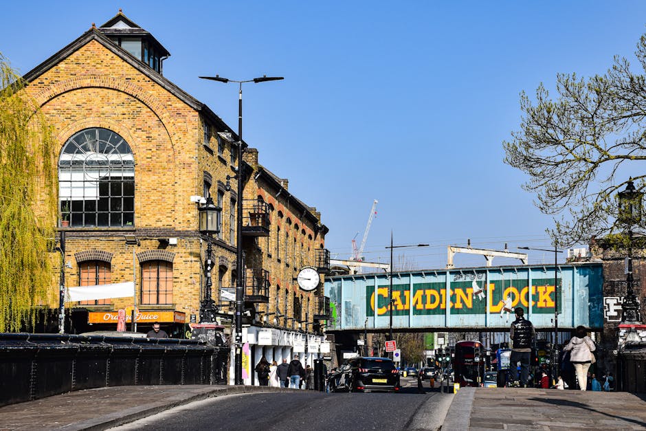 The image depicts a busy street scene in West Brompton with a clear blue sky overhead. A large, historic brick building with arched windows and multiple floors is visible on the left side. In front of