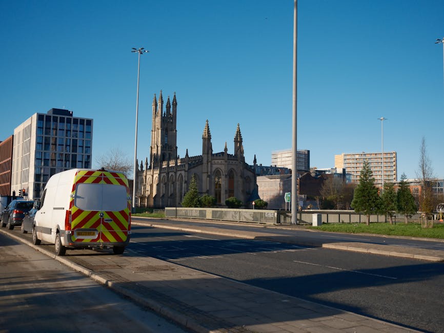 A white commercial van with red and yellow chevron reflective markings on its rear doors is parked on the side of a paved street in West Brompton, near a curb and a sidewalk. The van is positioned adjacent to a multi-story modern building with a grid-like facade, and behind it, there is a historic Gothic-style church with tall spires and ornate architectural details. In the foreground, there are three tall streetlights, and to the right, a few small trees and a grassy area are visible. The scene is captured under clear blue skies, indicating daytime, and the environment suggests an urban setting suitable for home relocation and furniture transport activities, with nearby buildings and infrastructure supporting moving logistics. The image relates to house removals and moving services provided by Man with Van West Brompton, illustrating the typical urban moving environment.