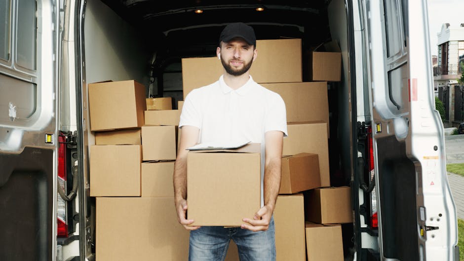 A man with a beard, dressed in a white polo shirt, jeans, and a black cap, stands at the open rear of a moving van on a residential street. Inside the van, numerous cardboard boxes of various sizes are stacked, some secured with plastic wrap. The man is holding a box close to his body, with a clipboard resting on top, indicating a packing or inventory process. The van is parked on the pavement near houses with visible doors and balconies. The image captures a home relocation scene, highlighting furniture transport and packing aspects of a moving process, with natural daylight illuminating the scene. Man with Van West Brompton provides professional removals services and uses equipment such as trolleys and blankets to assist in safe and efficient furniture transport during house moves.